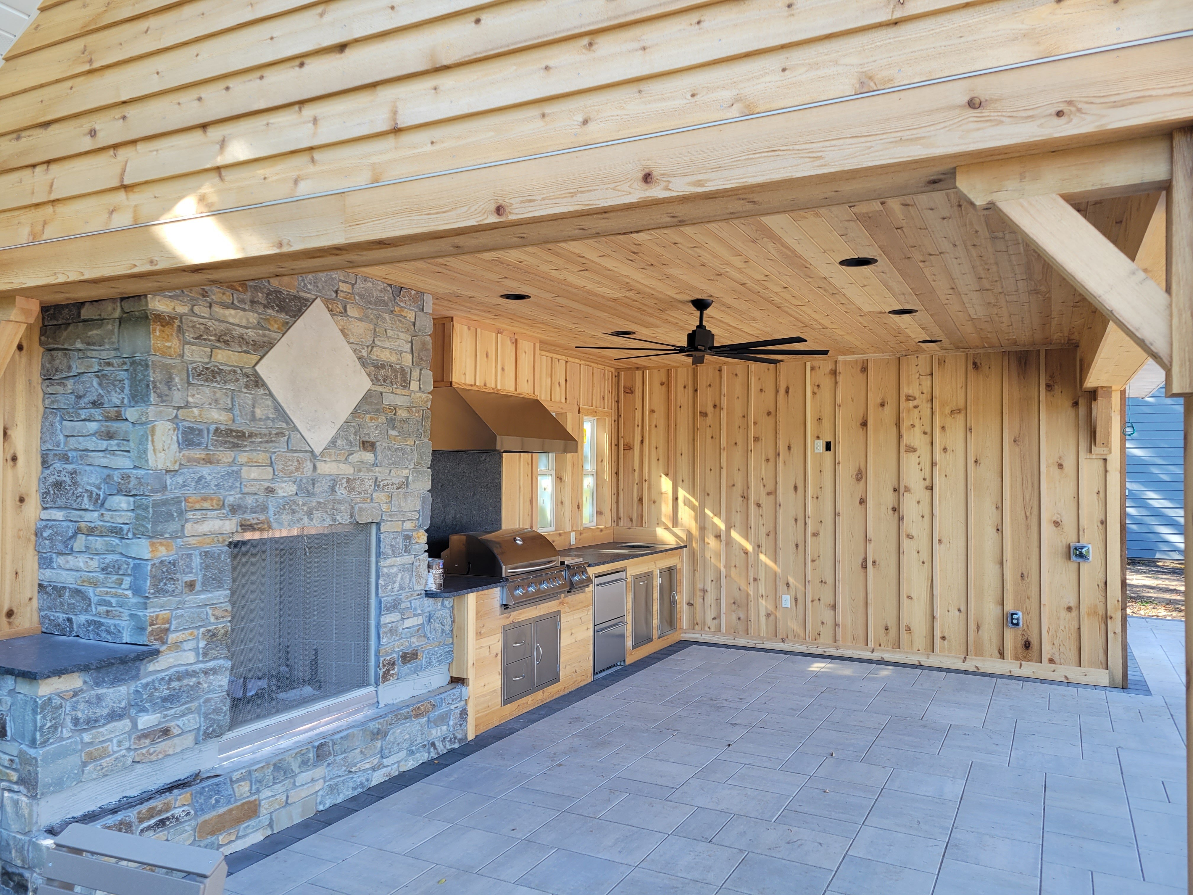 Covered outdoor kitchen with natural wood paneling, a large stone fireplace, built-in grill, stainless appliances, and ceiling fan. The structure features exposed beams, tile flooring, and custom finishes for a cozy, all-season outdoor living area.