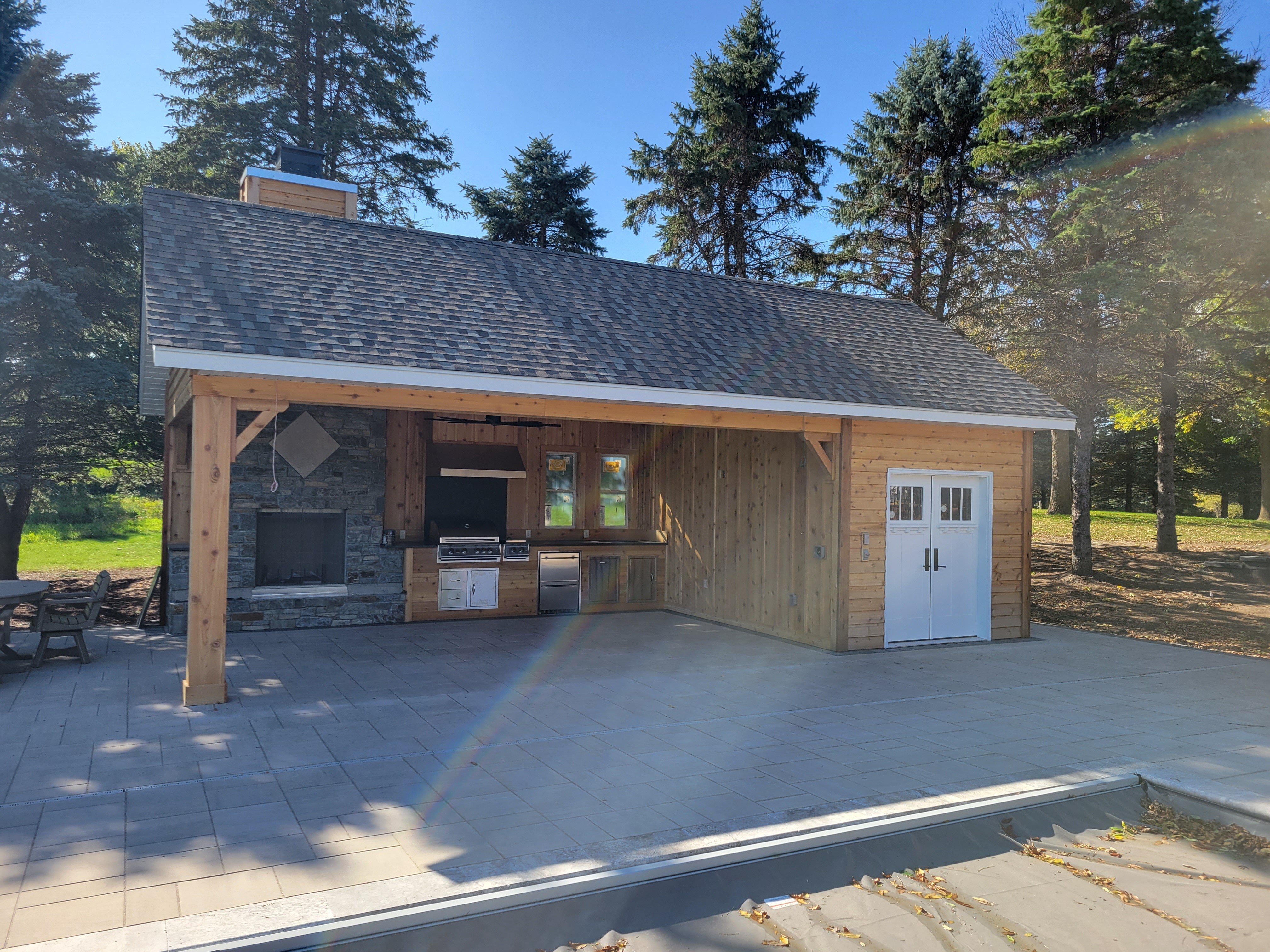 Free-standing structure with pitched roof, wood siding, and stone fireplace, housing a built-in grill and appliances on a paved patio. Positioned next to a pool with a clean, open layout.