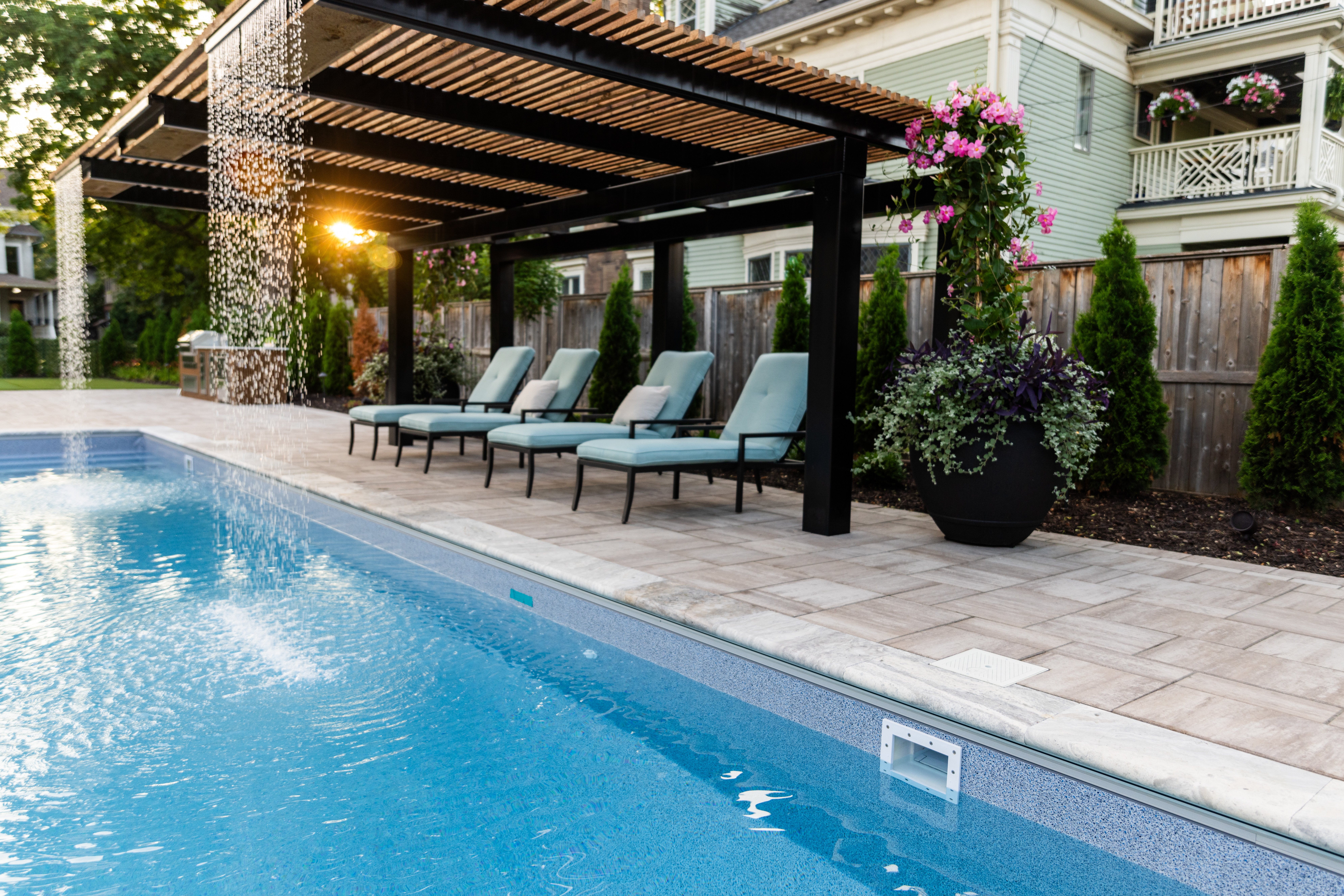 Four cushioned loungers beneath a black steel pergola with wooden slats and cascading water features beside a pool. Paver deck, flowering planters, and privacy landscaping complete the serene setup.
