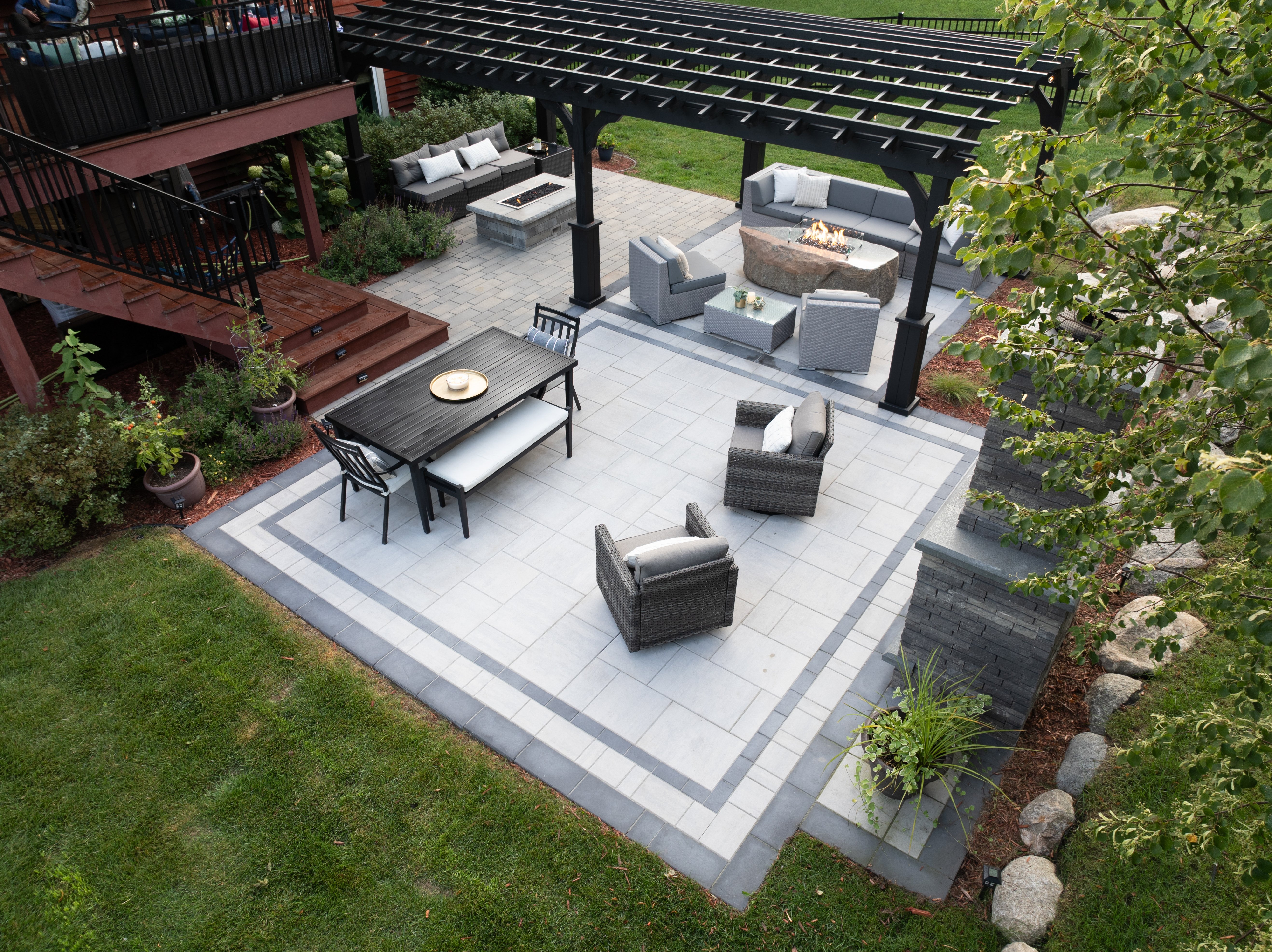 Spacious backyard patio divided into lounge and dining zones with gray pavers, black pergola, fire pit, and modern outdoor furniture. Tiered layout connects to a wood deck with stairs and surrounding greenery.