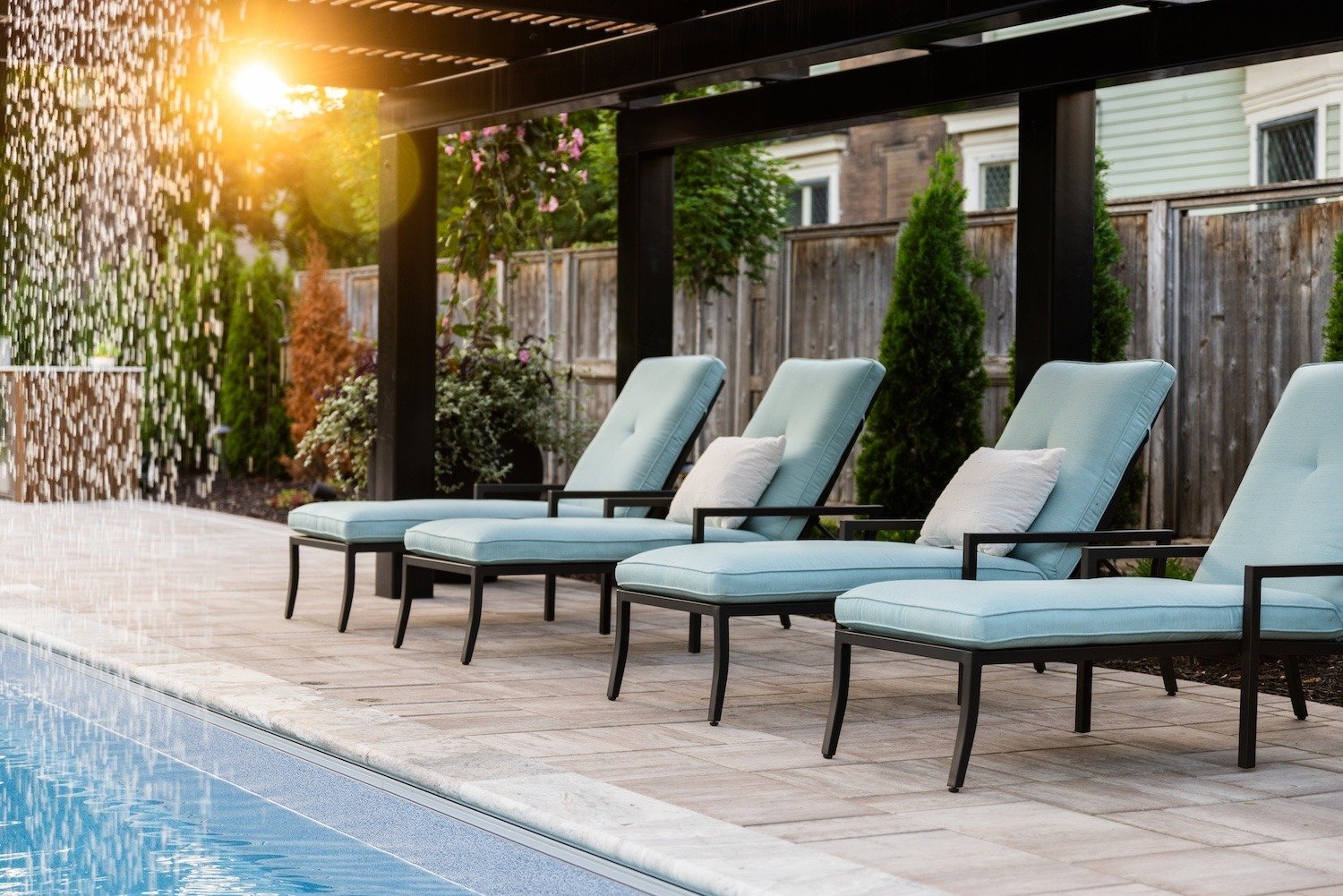 an in-ground pool surrounded by a paver and lounge chairs. above the chairs is a pergola. An attached waterfall creates a beautiful water display of trickling water from the pergola into the pool.