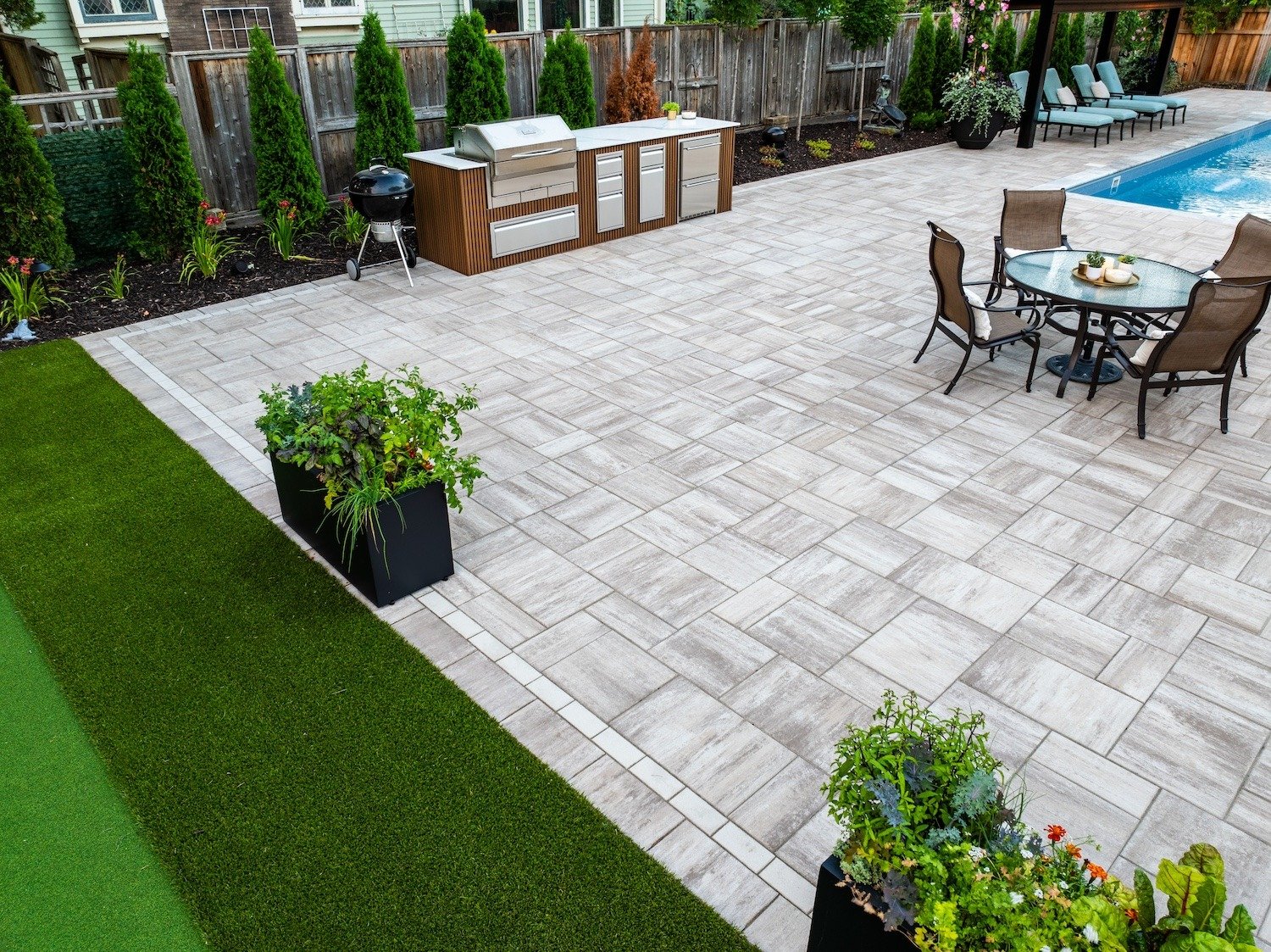 backyard with a large hardscaped paver surrounded by green grass. an outdoor grill is nearby, alongside an outdoor dining table with chairs.