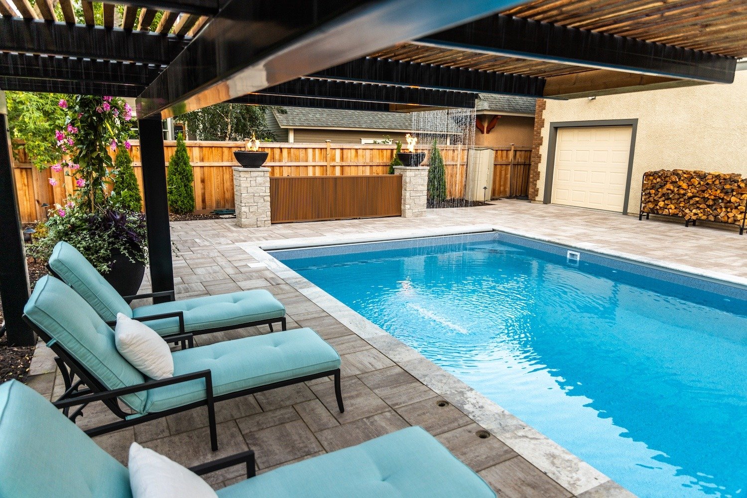 an in-ground pool surrounded by a paver and lounge chairs. above the chairs is a pergola. An attached waterfall creates a beautiful water display of trickling water from the pergola into the pool.