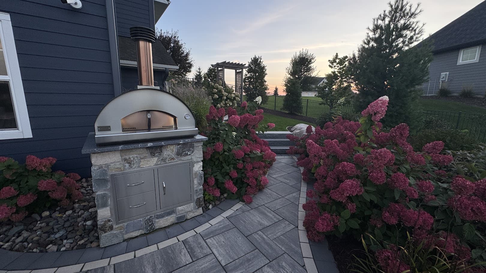 a flower garden in a backyard. lush, blooming pink flowers surrounding a path with a nearby pizza oven.