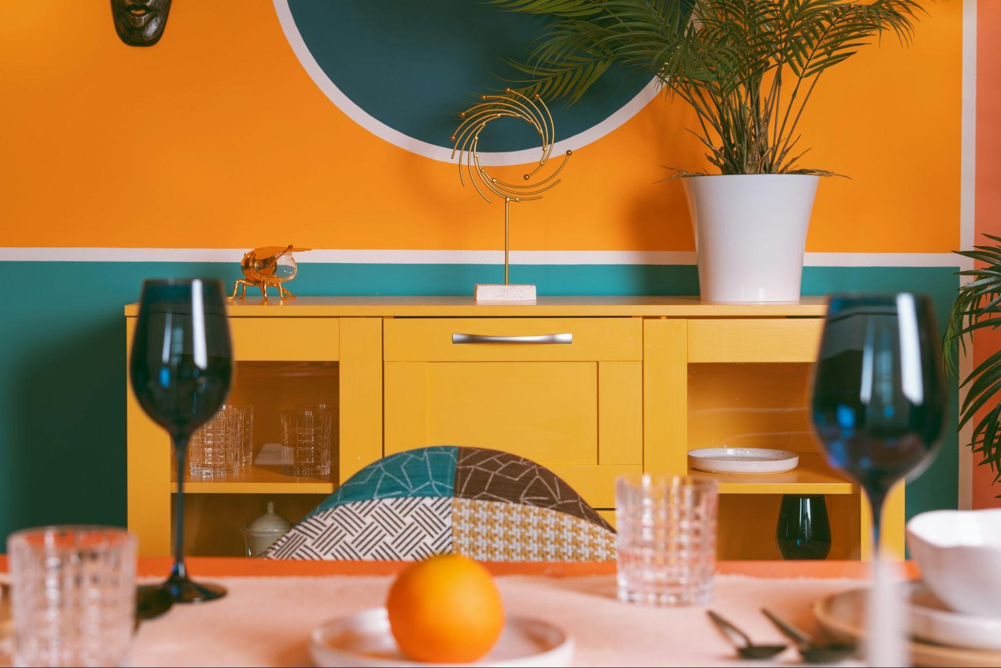 bright and colorful dining room with yellow cabinet, orange and blue wall, and colorfully patterned chairs.