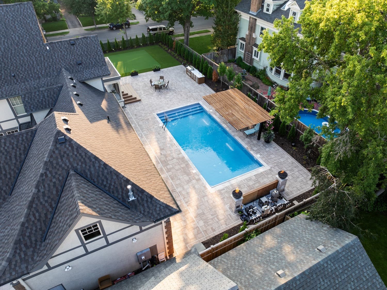 an in-ground pool in a compact backyard, with surrounding pavers, a pergula, and an outdoor kitchen