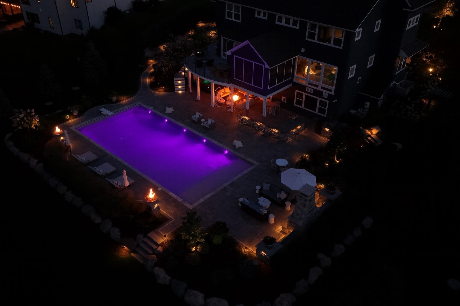 a backyard with an in-ground pool at night. Multiple fire features provide lighting: a fire pit, several fire bowls, and an outdoor fireplace