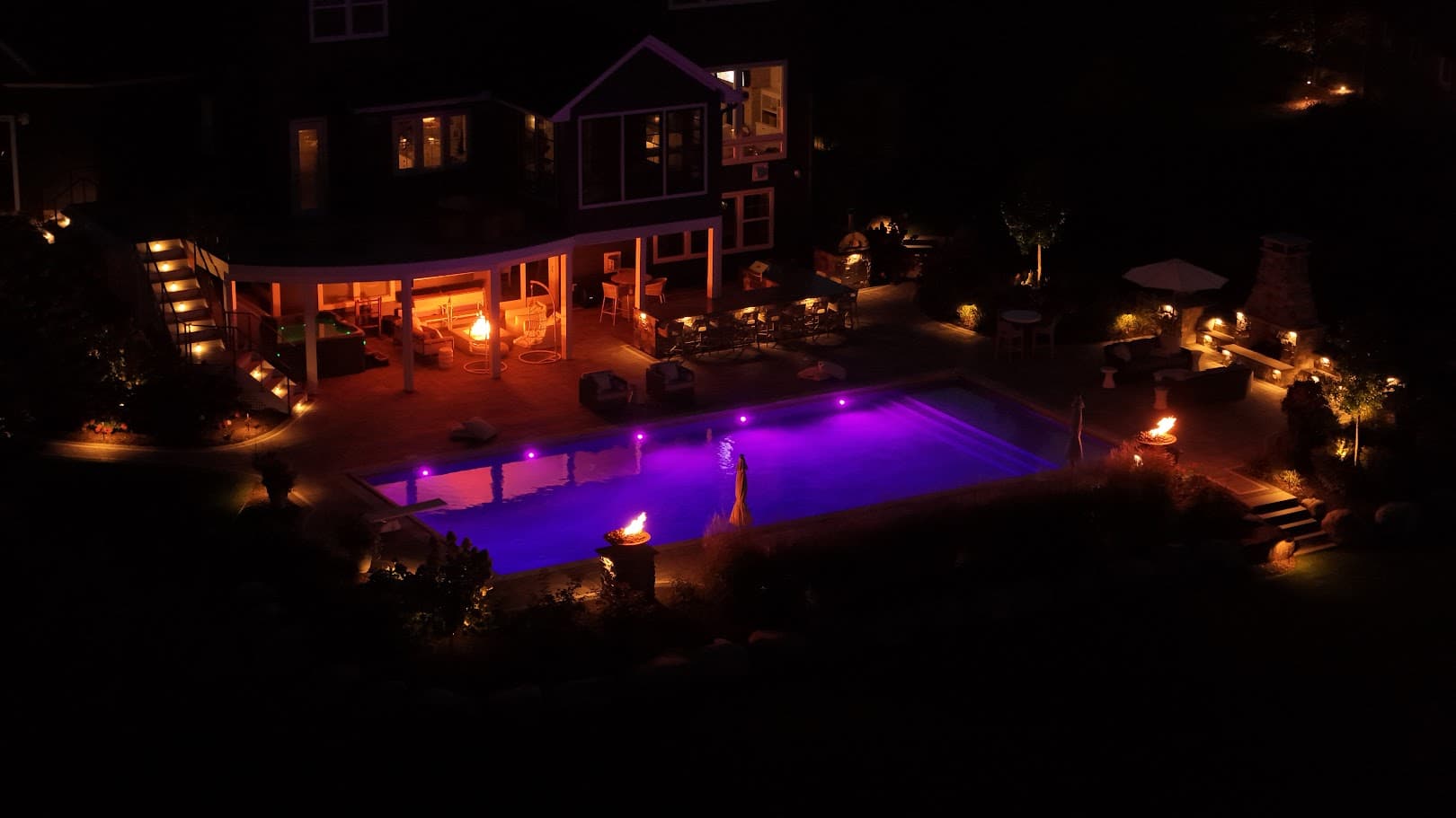 a backyard with an in-ground pool at night. Multiple fire features provide lighting: a fire pit, several fire bowls, and an outdoor fireplace