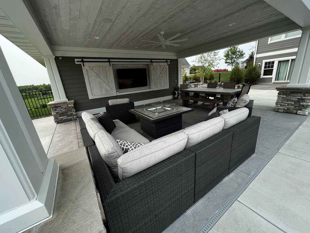 a partially covered outdoor living room with sectional seating, TV, dining table, and fire pit 