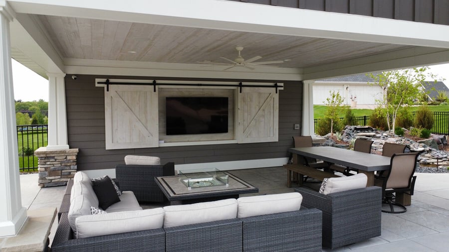 an outdoor living room. a TV rests against the wall of a poolhouse, with barn-style doors that provide protection. around the VT is a modern fire table surrounded by a large outdoor sectional.