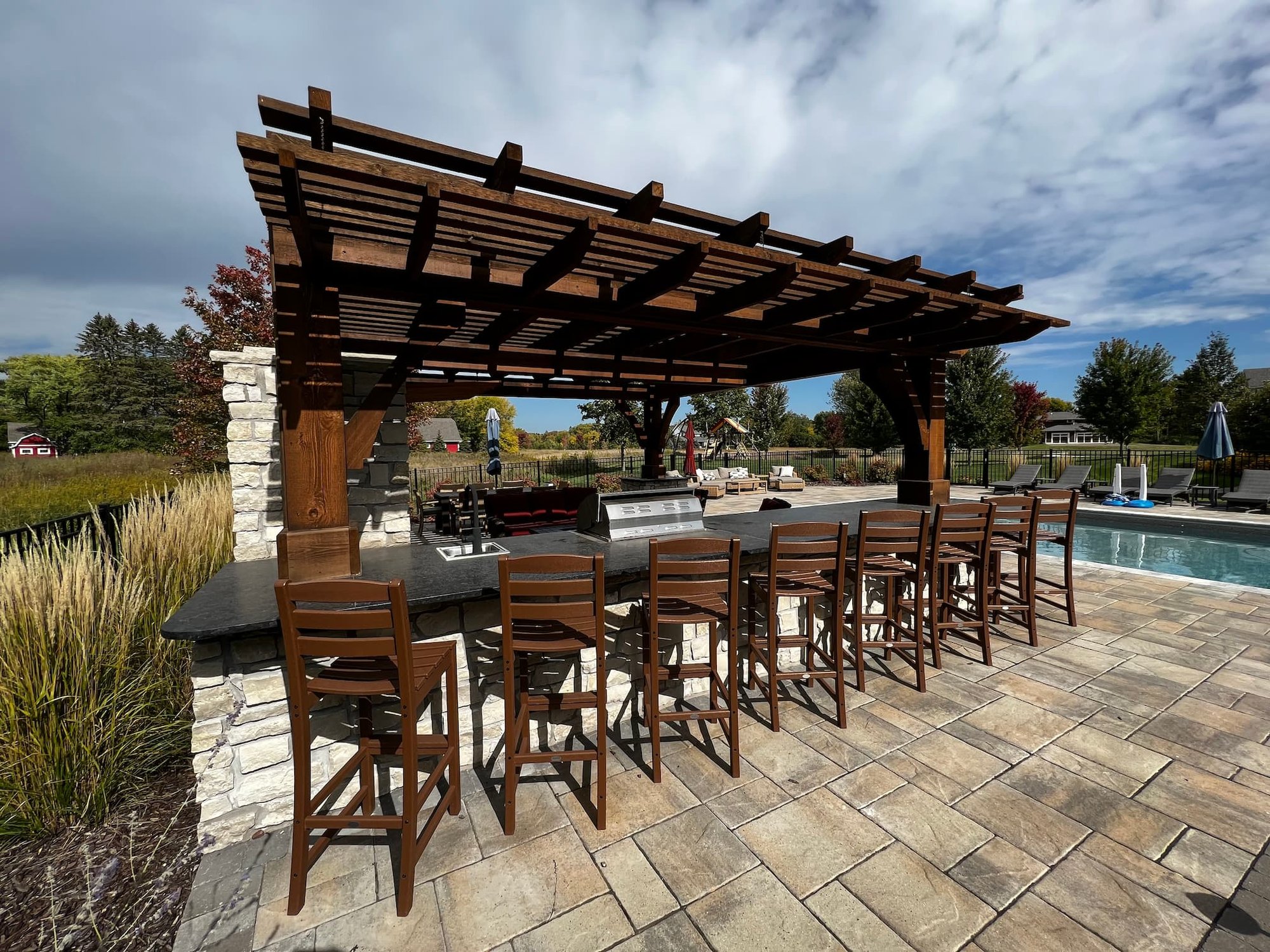 a bar area in an outdoor kitchen. a beautiful dark wood pergola partiall covers a custom black countertop, which is flanked by 8 bar chairs.