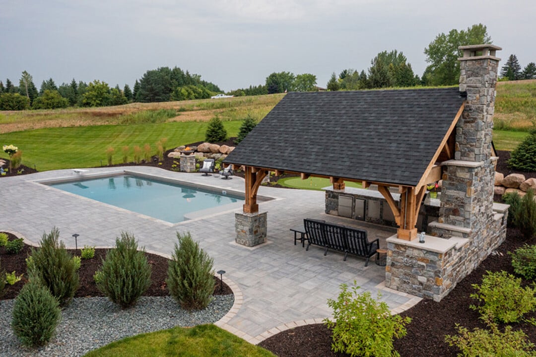a pool surrounded by a beautiful paver patio. A stone and wood pergola with an outdoor kitchen