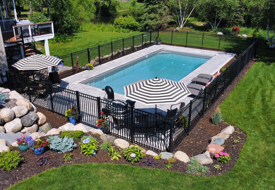 Rectangular in-ground pool surrounded by a black metal safety fence, lounge chairs, and striped patio umbrellas. The landscape features colorful flower beds, stone edging, and a backdrop of trees and lawn—combining pool design with vibrant garden elements.