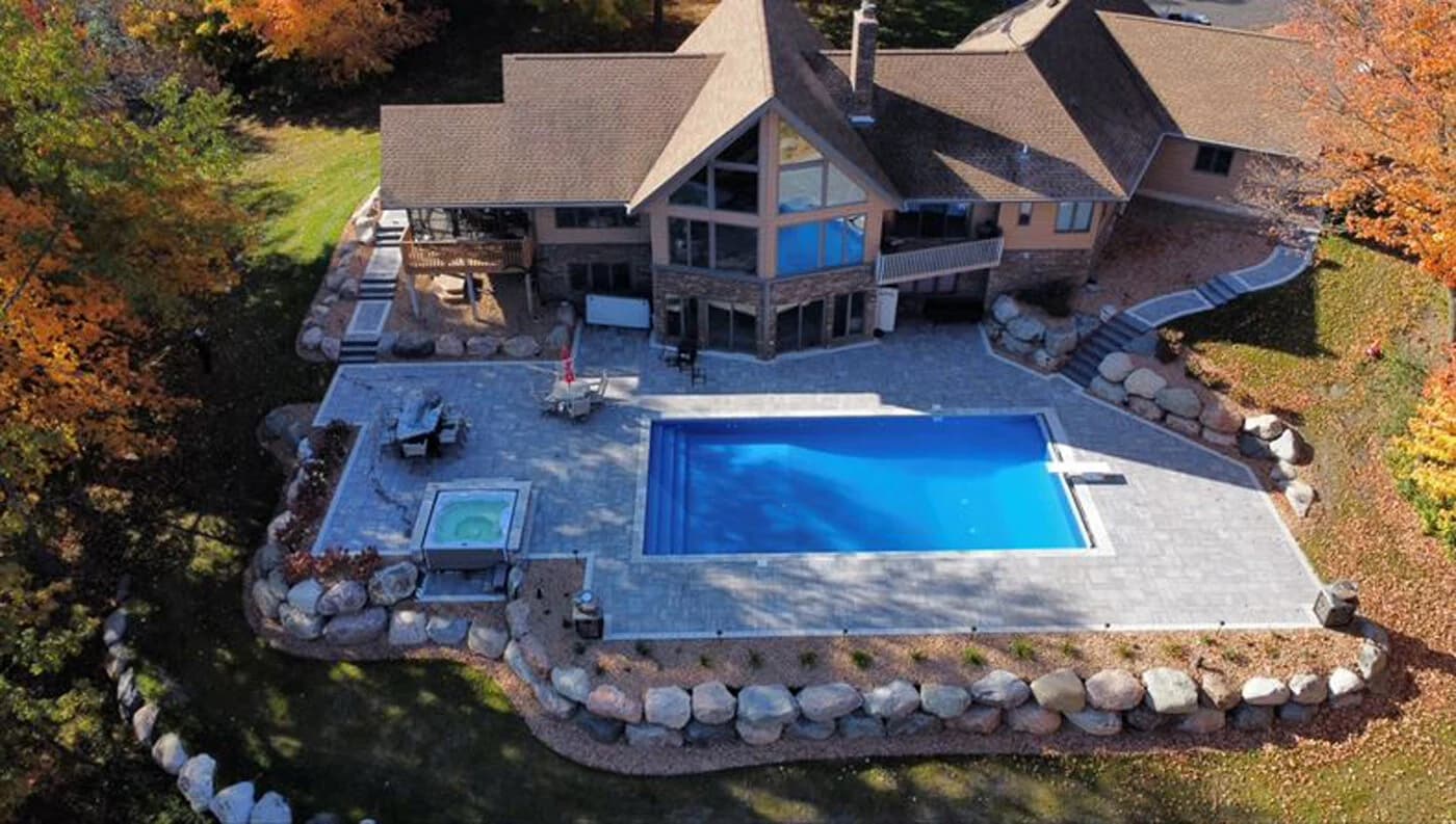 an aerial shot of a backyard with a large pool and hottub surrounded by a paver and large retaining wall.