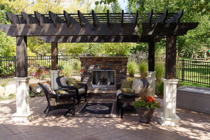 an outdoor fireplace surrounded by chairs. a wooden pergola provides some shade.