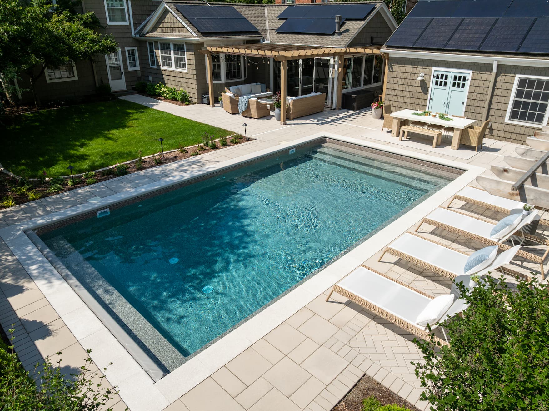 an in ground pool in a backyard, surrounded by gorgeous pavers, an outdoor living room, a pool house, and lounge chairs.