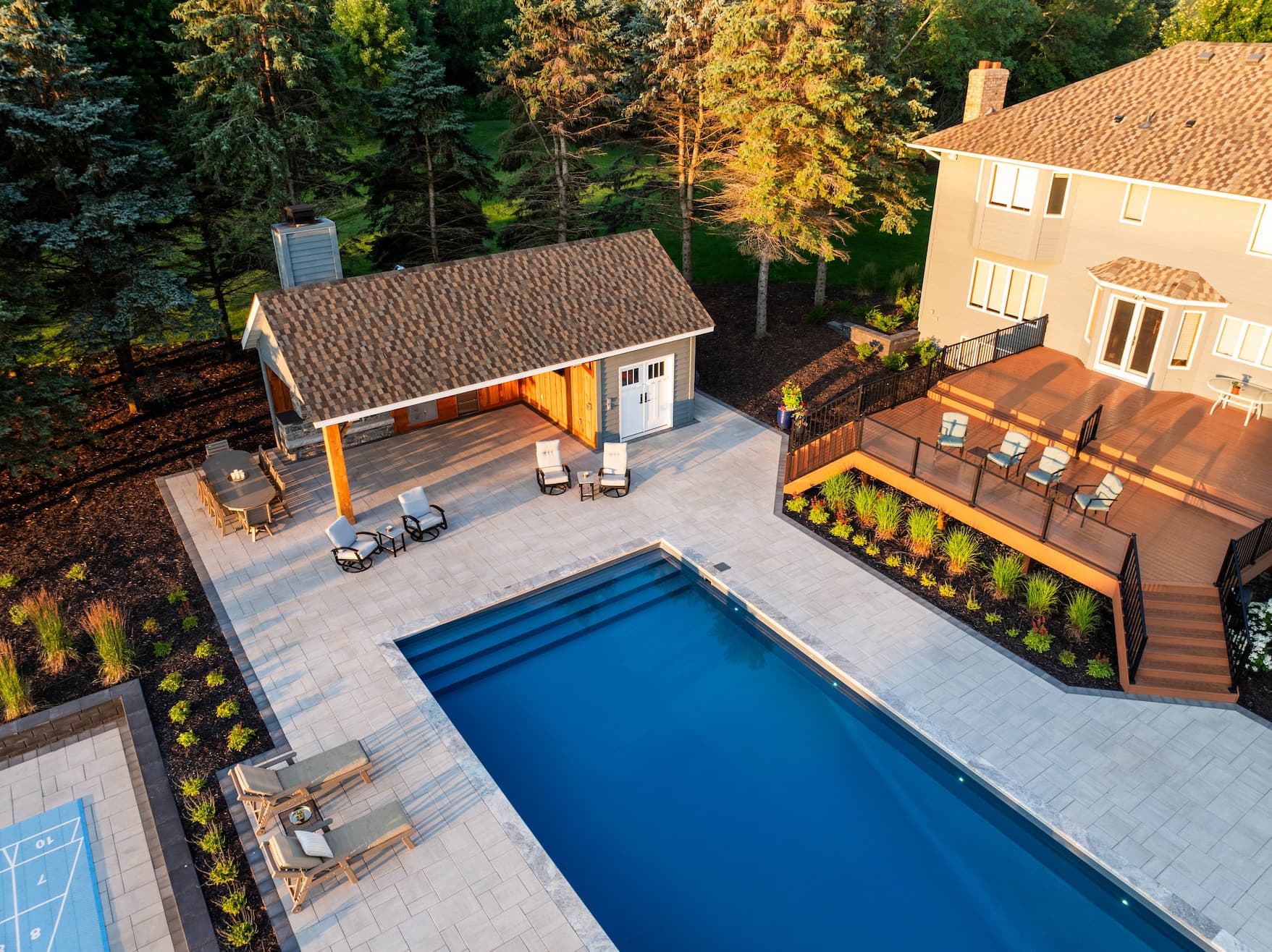a backyard with beautiful in-ground pool surrounded by gray pavers. a shuffleboard and pool house are nearby.