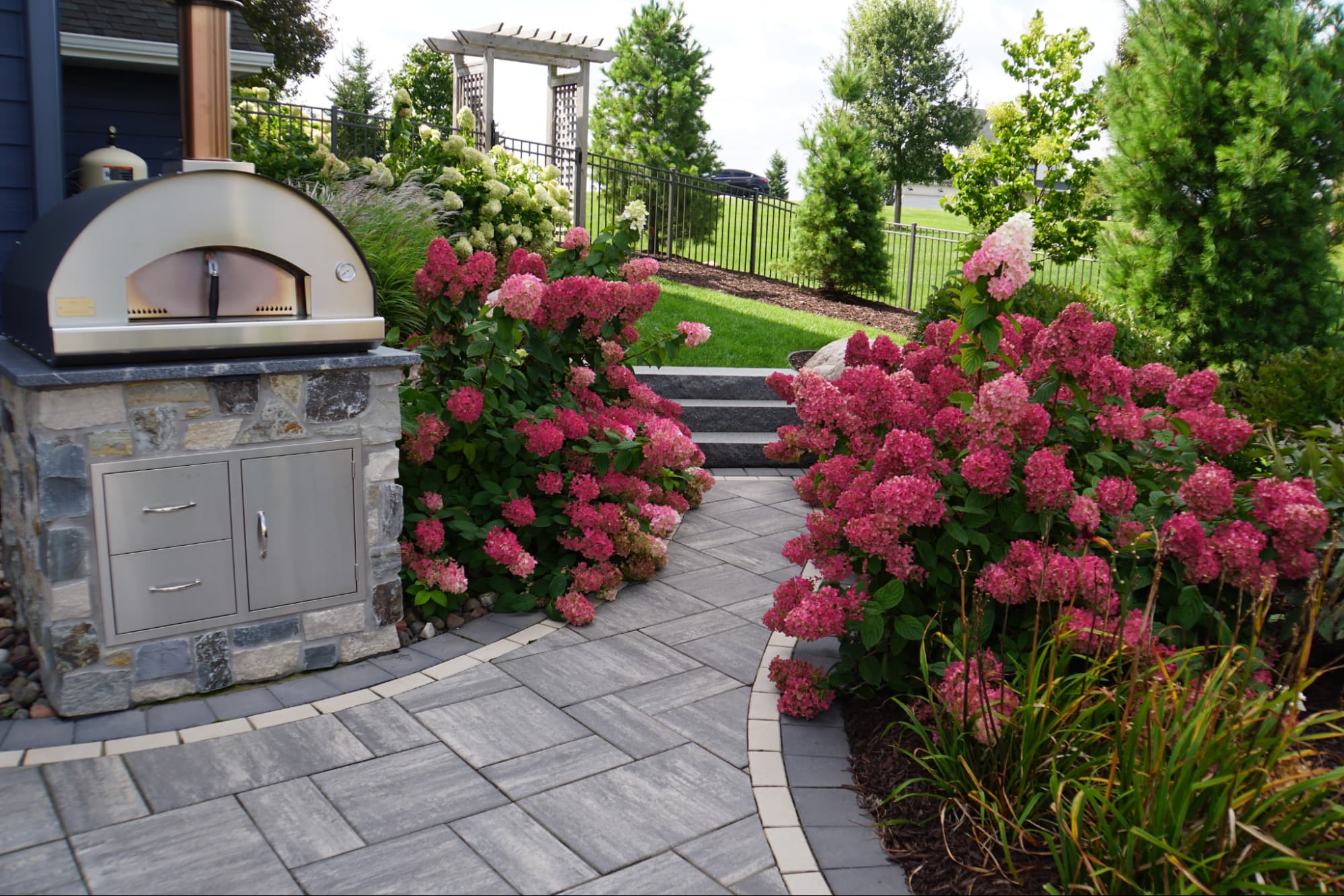 a paved pathway leading through a garden area of a backyard. on one side is a bespoke pizza oven. on the other are blooming pink flowers.