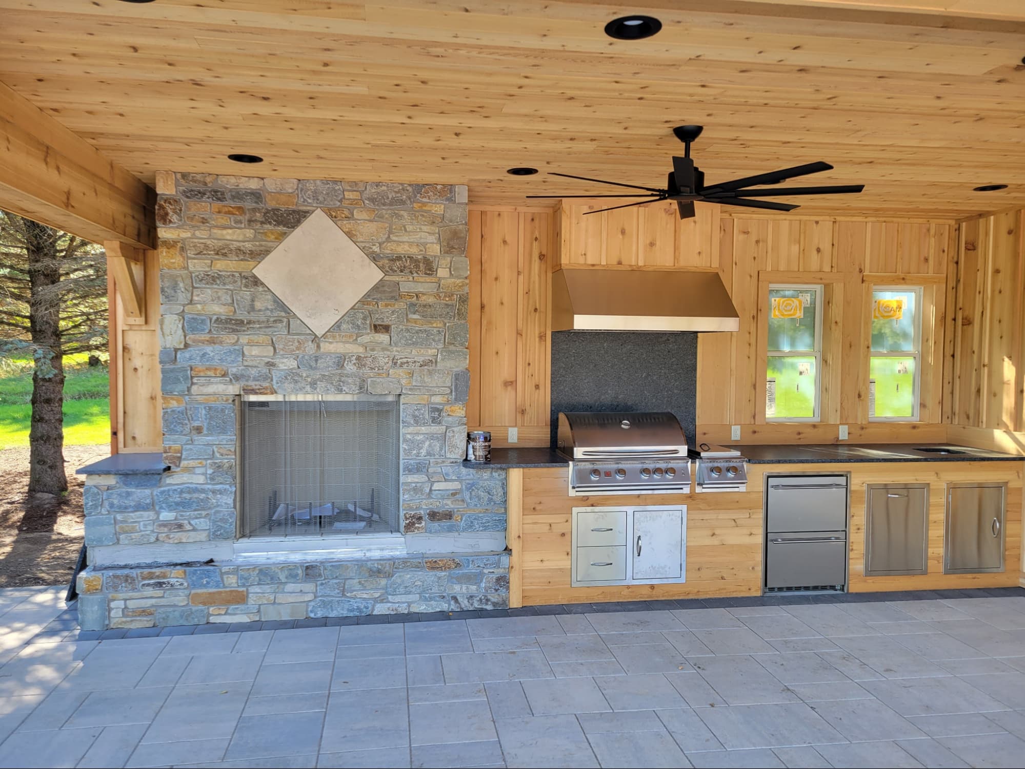Covered outdoor kitchen with knotty pine walls and ceiling, featuring a built-in stone fireplace, stainless grill, ventilation hood, mini fridge, and sink. A functional and stylish outdoor living space with custom wood and stone finishes.